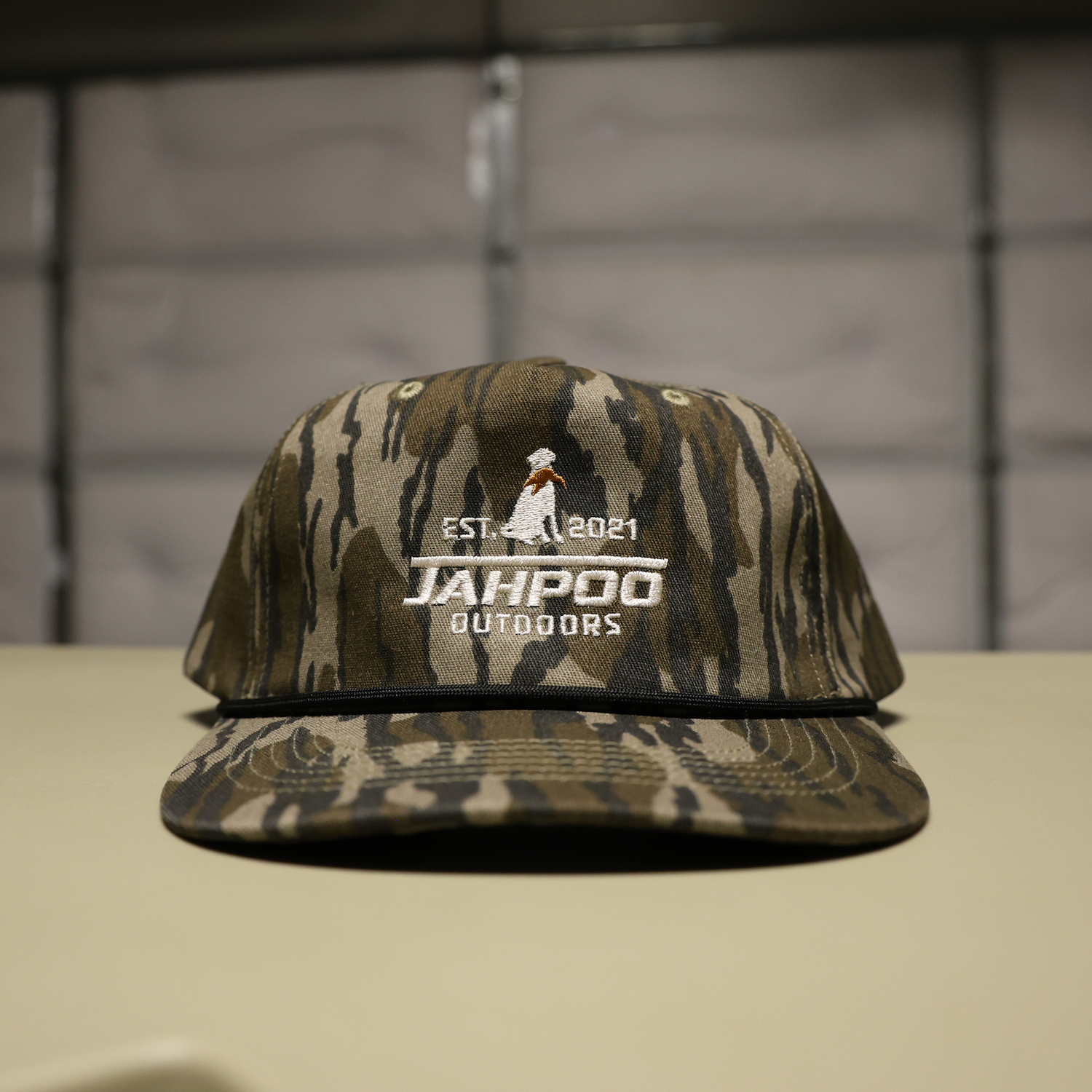 A camouflage baseball cap with a white embroidered logo reading 'JAHPOO OUTDOORS' and 'EST. 2021' sits on a light-colored surface against a dark, blurred background.