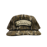 A camouflage baseball cap with a prominent 'JAHPOO OUTDOORS' patch on the front, displayed on a beige surface against a gray brick wall background.