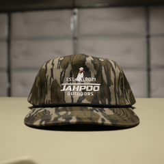 A camouflage baseball cap with a white embroidered logo reading 'JAHPOO OUTDOORS' and 'EST. 2021' sits on a light-colored surface against a dark, blurred background.