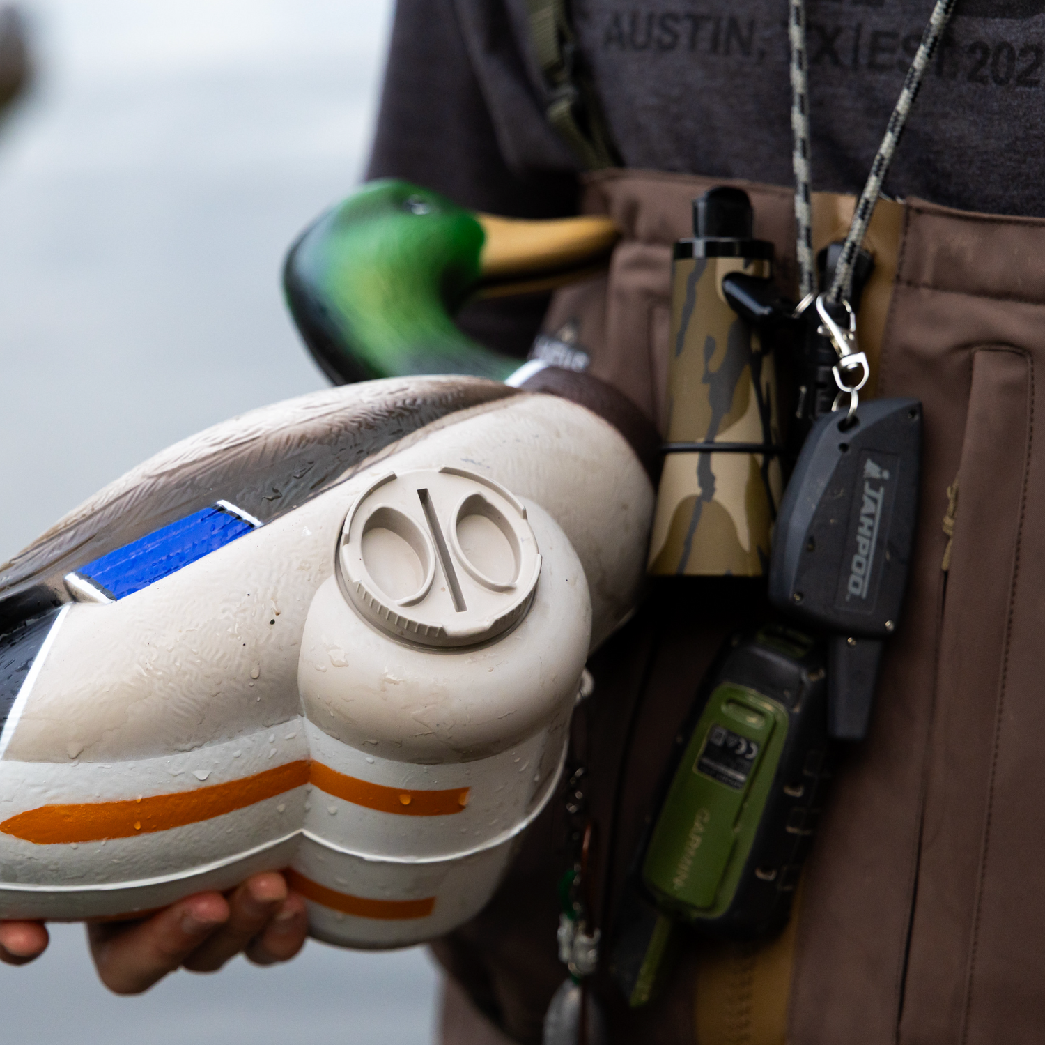 A person in a brown waders vest holds a realistic duck hunting deco shaped like a mallard duck head, with another duck deco and hunting accessories hanging from the vest against a blurred water background.